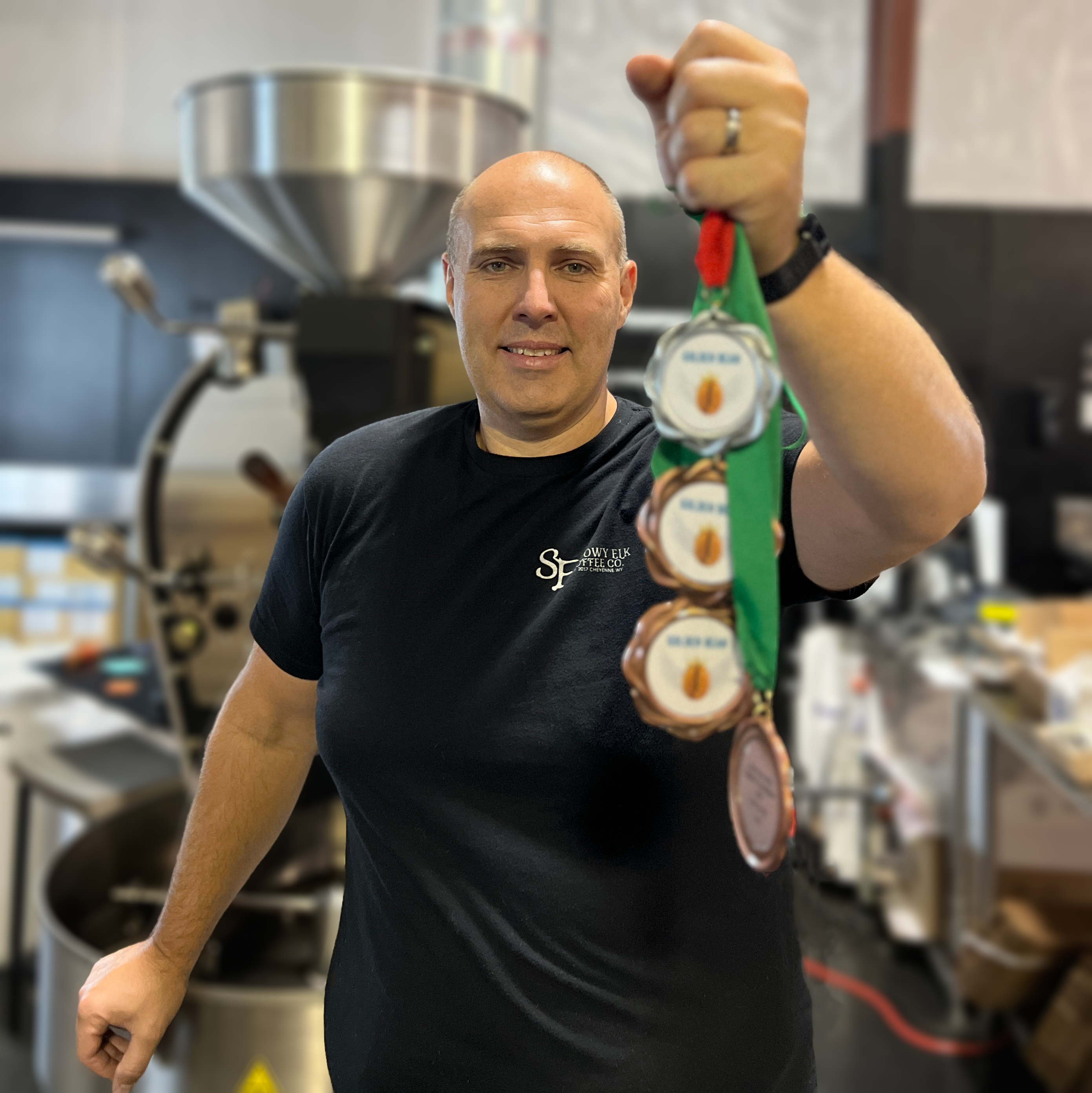 A man standing in front of a coffee roaster holding 4 medals