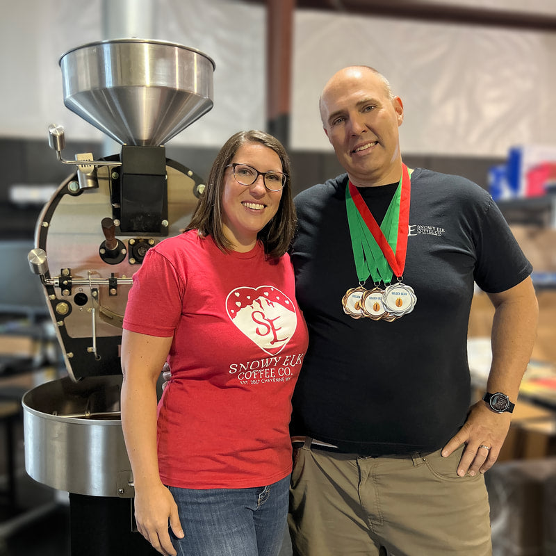 A couple standing near an industrial coffee roaster. The man has several medals around his neck.