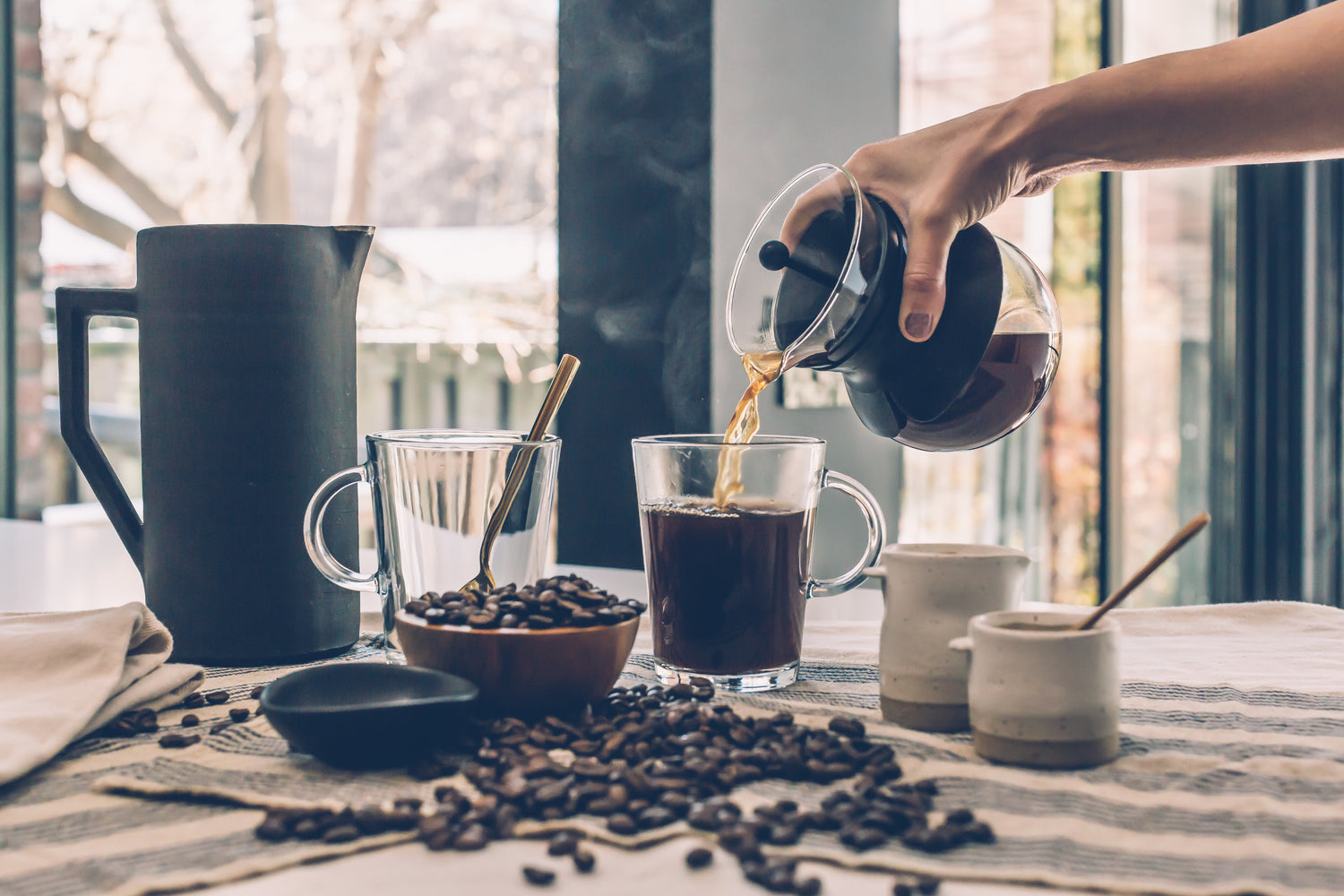 Pouring hot coffee from a pour over carafe surrounded by coffee beans, cups, and a large black kettle