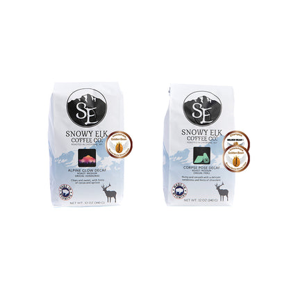 Two packages of Snowy Elk coffee, one labeled 'Alpine Glow Decaf' and the other 'Corpse Pose Decaf', each in a white bag with product name and an image of a deer.