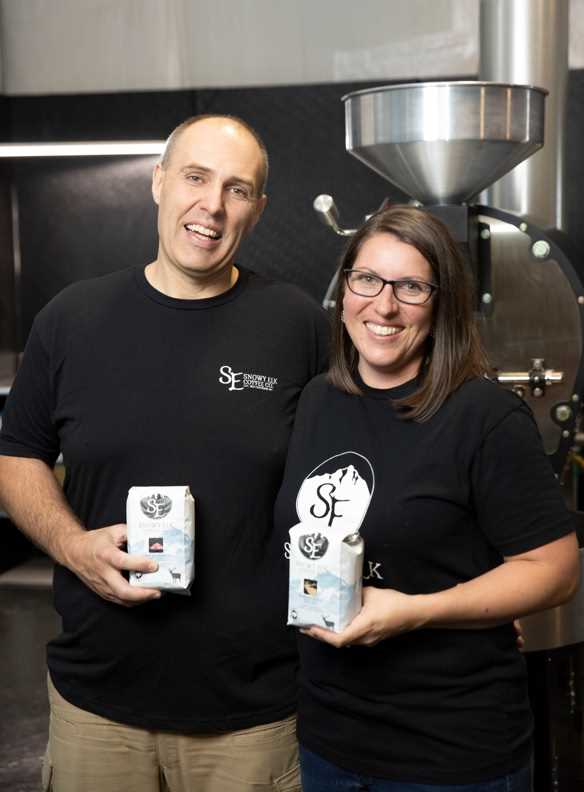 A couple wearing black t shirts holding bags of coffee in front of an industrial coffee roaster