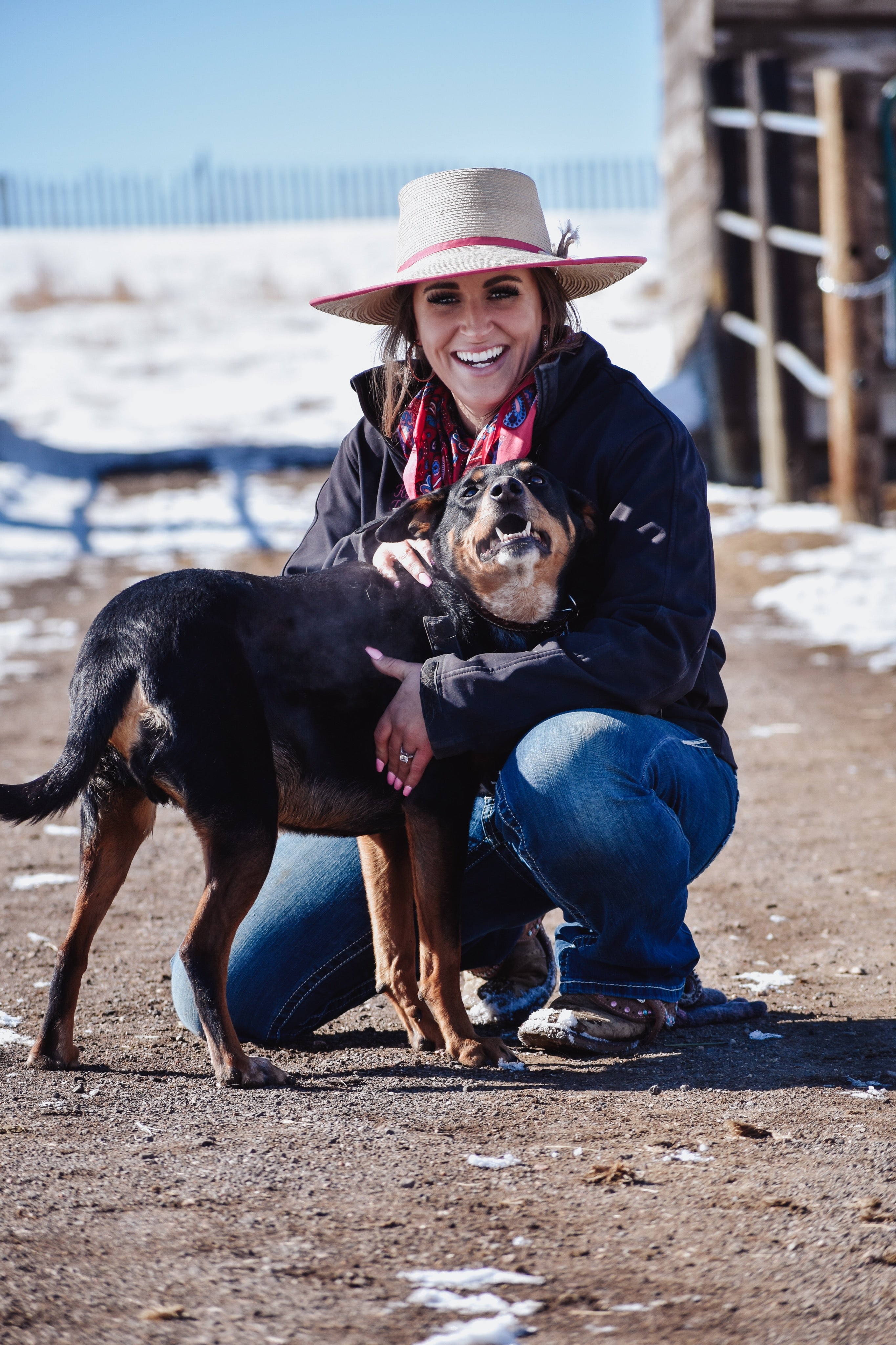 Woman in a cowboy hat and scarf holding a dog outdoors on a sunny day.