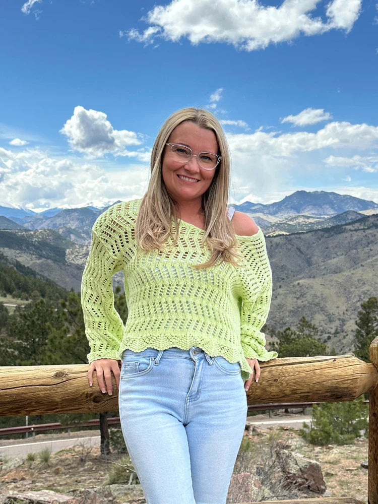 A woman with blonde hair and glasses wearing a green shirt with a mountain background