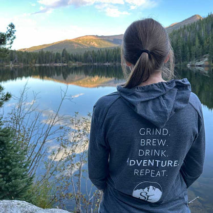 A woman with dark hair standing near a lake wearing a Snowy Elk shirt that says Grind. Brew. Adventure. Repeat.