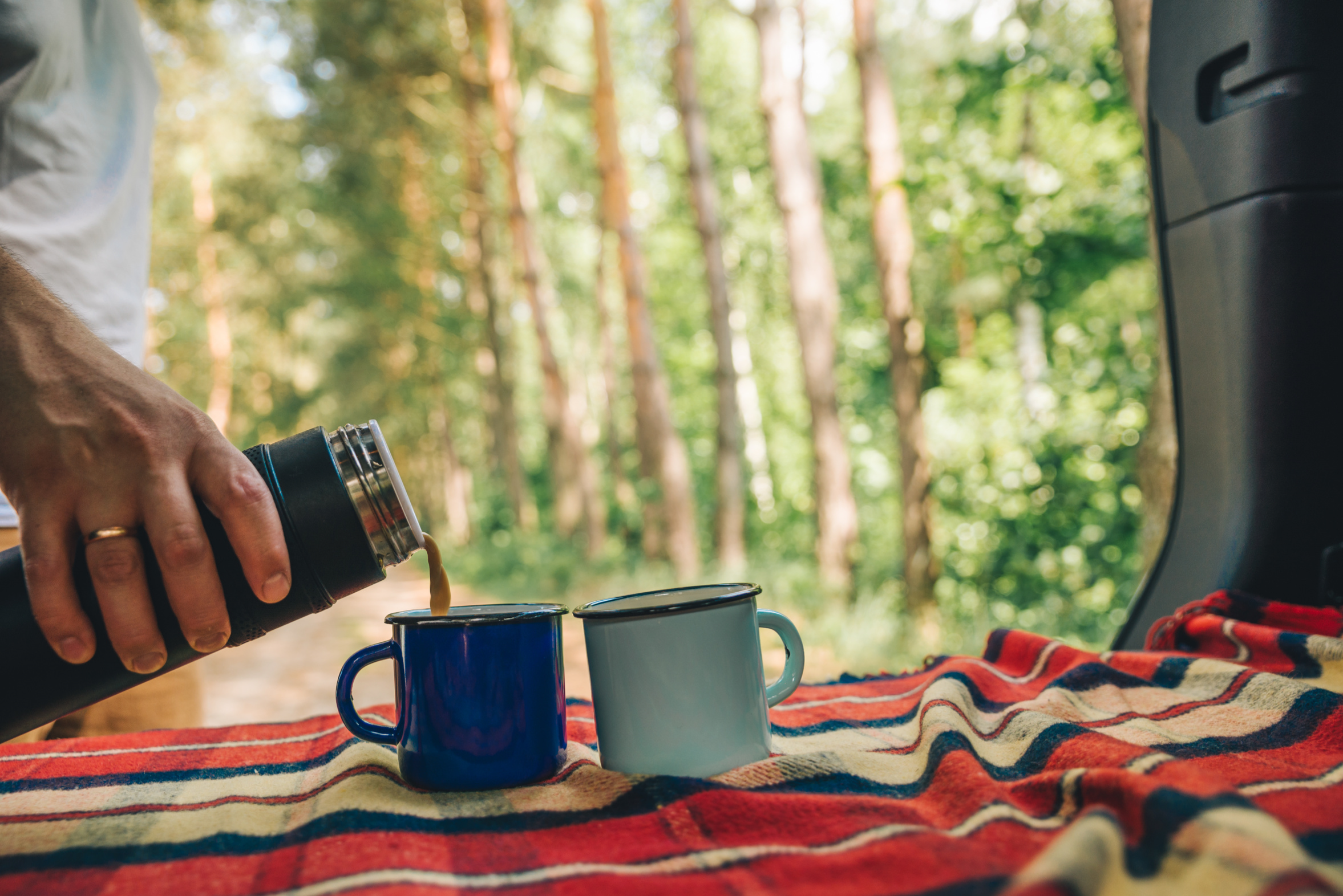 Person pouring coffee from a thermos into two mugs on a camping mat with a forest background